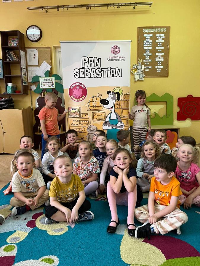 grupa dzieci siedząca w kucki na dywanie nad nimi plakat z napisem pan sebastian