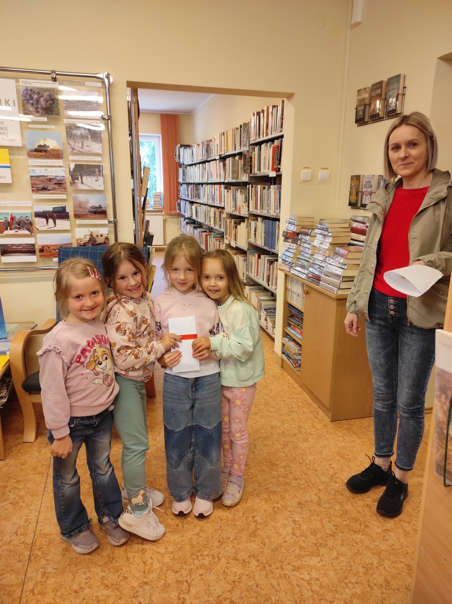po prawej stronie stoi kobieta z kartką w ręku po lewej czwórka małych dziewczynek trzyma białą kopertę w tle widzoczne regały z książkami 