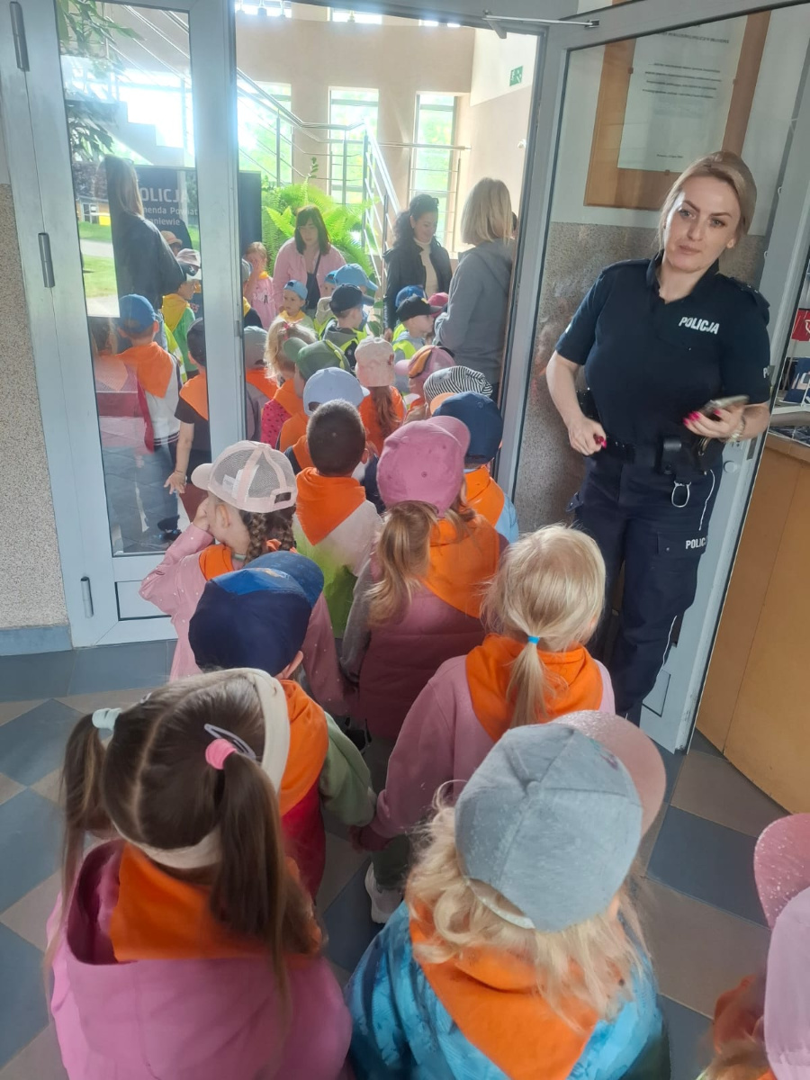 Grupa dzieci w pomarańczowych chustach stoi w korytarzu komendy policji. W tle widoczne są opiekunki a obok drzwi stoi policjantka w mundurze