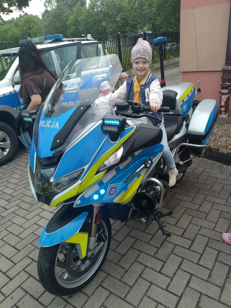 Uśmiechnięta dziewczynka w żółtej chuście siedzi na policyjnym motocyklu. Za nią widać radiowóz oraz inne dzieci oglądające pojazdy policyjne