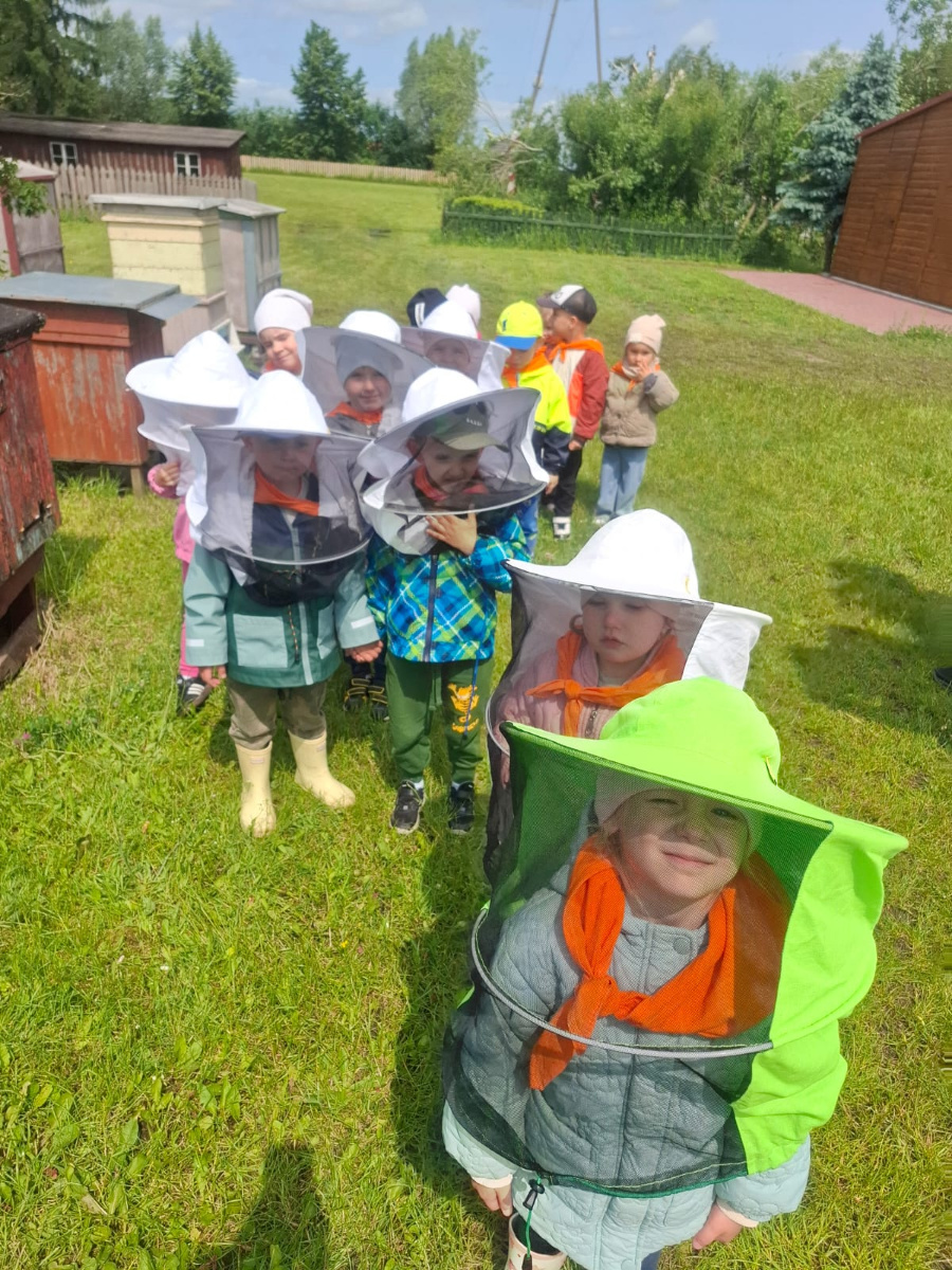 Grupa dzieci stoi na trawie ubrana w ochronne stroje pszczelarskie z siatkowanymi kapeluszami. Niektóre dzieci mają na sobie kolorowe kurtki i pomarańczowe chusty. W tle widoczne są ule