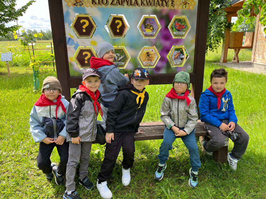 Sześciu chłopców siedzi na ławce przed tablicą edukacyjną z napisem Kto zapyla kwiaty. Dzieci mają kolorowe kurtki czapki i czerwone lub żółte chusty na szyjach