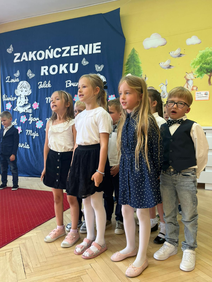 Grupa dzieci stoi w sali przed granatową dekoracją z napisem Zakończenie roku. Dziewczynki ubrane są w białe bluzki i ciemne spódnice a chłopiec obok nich ma koszulę kamizelkę i muszkę