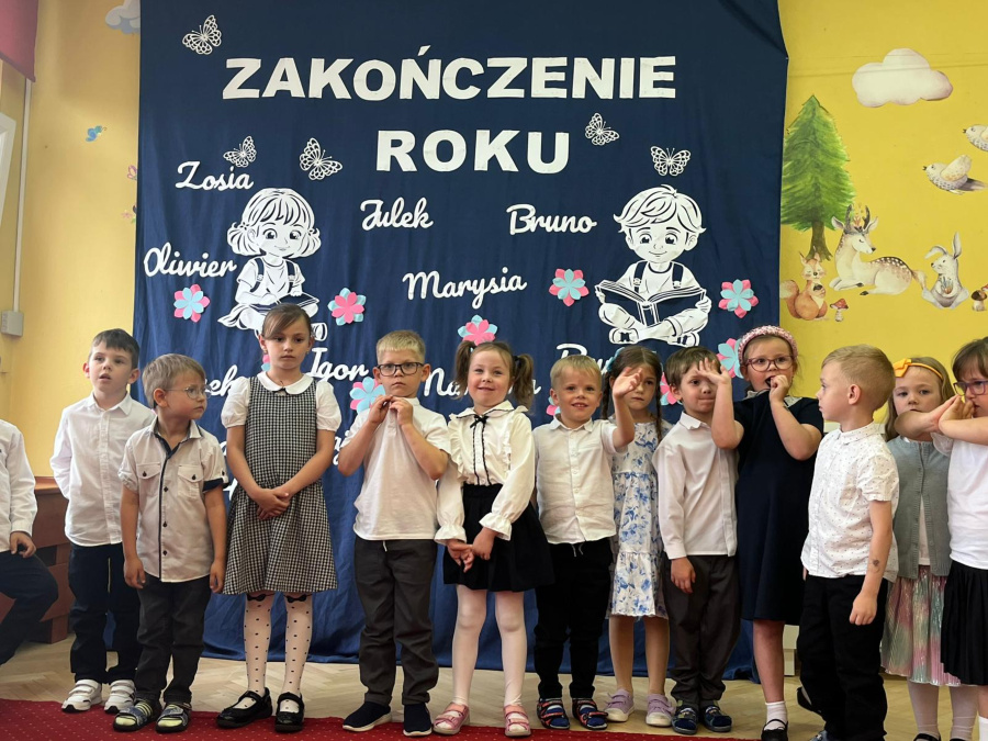 Grupa dzieci ubranych odświętnie stoi na tle granatowej dekoracji z napisem Zakończenie roku. Dzieci ustawione są w szeregu