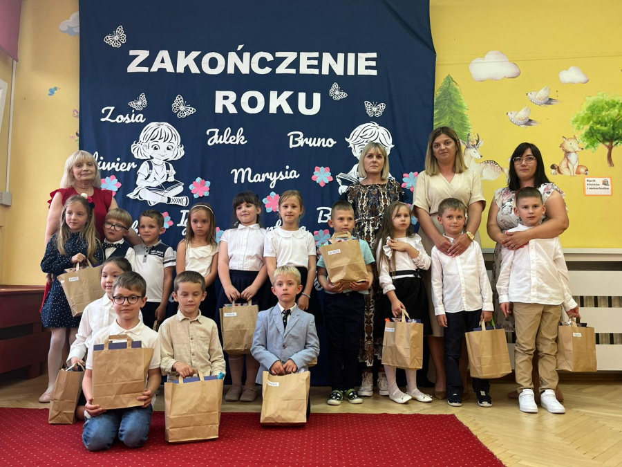 Grupa dzieci wraz z nauczycielkami pozuje do pamiątkowego zdjęcia na tle granatowej dekoracji z napisem „Zakończenie roku”. Dzieci ubrane są w białe koszule, sukienki i eleganckie spódnice