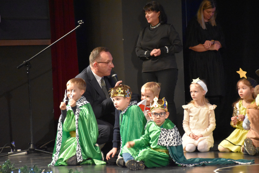 Na pierwszym planie troje dzieci przebranych za króli w zielonych pelerynach i koronach siedzi lub klęczy na scenie. Obok nich klęczy mężczyzna w garniturze z mikrofonem