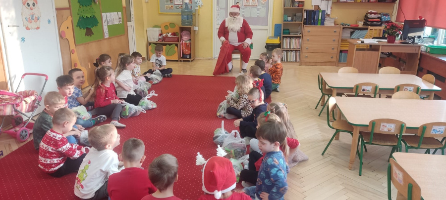 Dzieci siedzą w kole na czerwonym dywanie w trzymając paczki z upominkami. Na wprost nich przy drzwiach siedzi Mikołaj w czerwonym stroju i z białą brodą. 