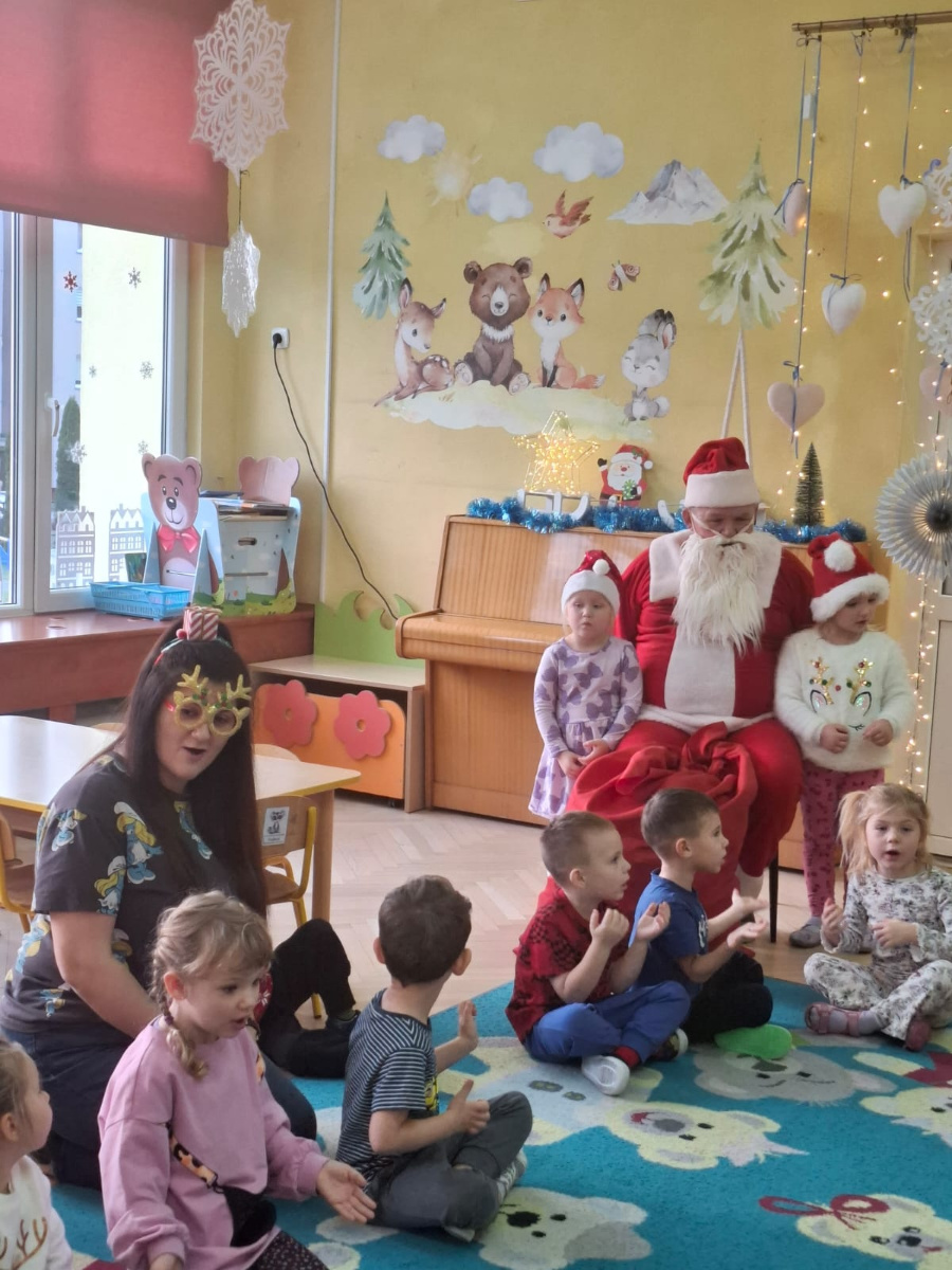 Na czerwonym dywanie siedzą dzieci wśród nich znajduje się Mikołaj w czerwonym stroju i z białą brodą. Niektóre dzieci mają na sobie świąteczne akcenty jak czapki Mikołaja czy opaski