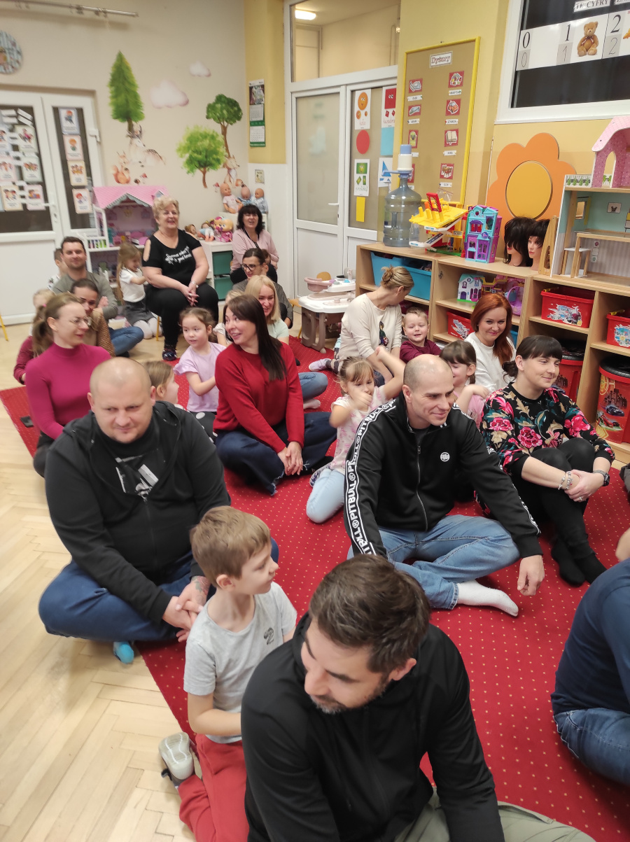 Dzieci i dorośli siedzą parami na czerwonym dywanie w sali przedszkolnej ustawieni w rzędach jeden za drugim. W tle widać nauczycielki kolorowe regały z zabawkami oraz dekoracje ścienne przedsta