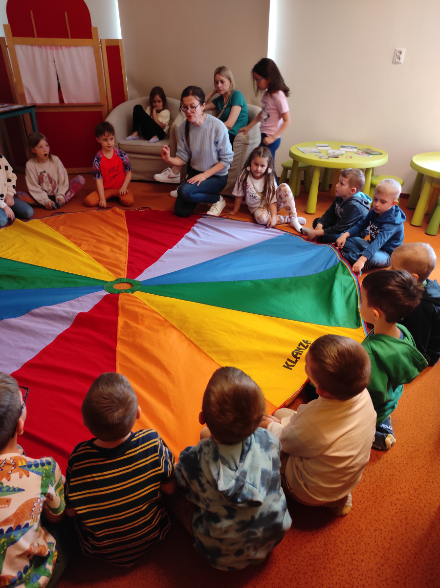 Dzieci siedzą w kręgu wokół dużej kolorowej chusty animacyjnej złożonej z barwnych trójkątów. Wspólnie trzymają jej brzegi. W tle widać stoliki krzesła i kilka osób