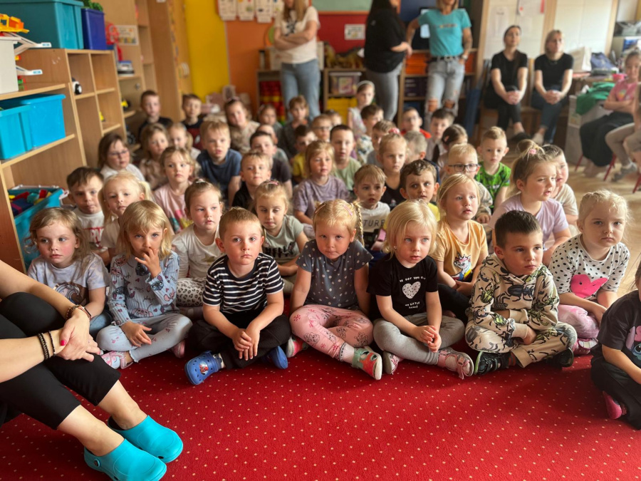 Duża grupa dzieci siedzi na czerwonym dywanie. Przedszkolaki uważnie patrzą przed siebie jakby słuchały występu lub oglądały przedstawienie. W tle stoją nauczycielki