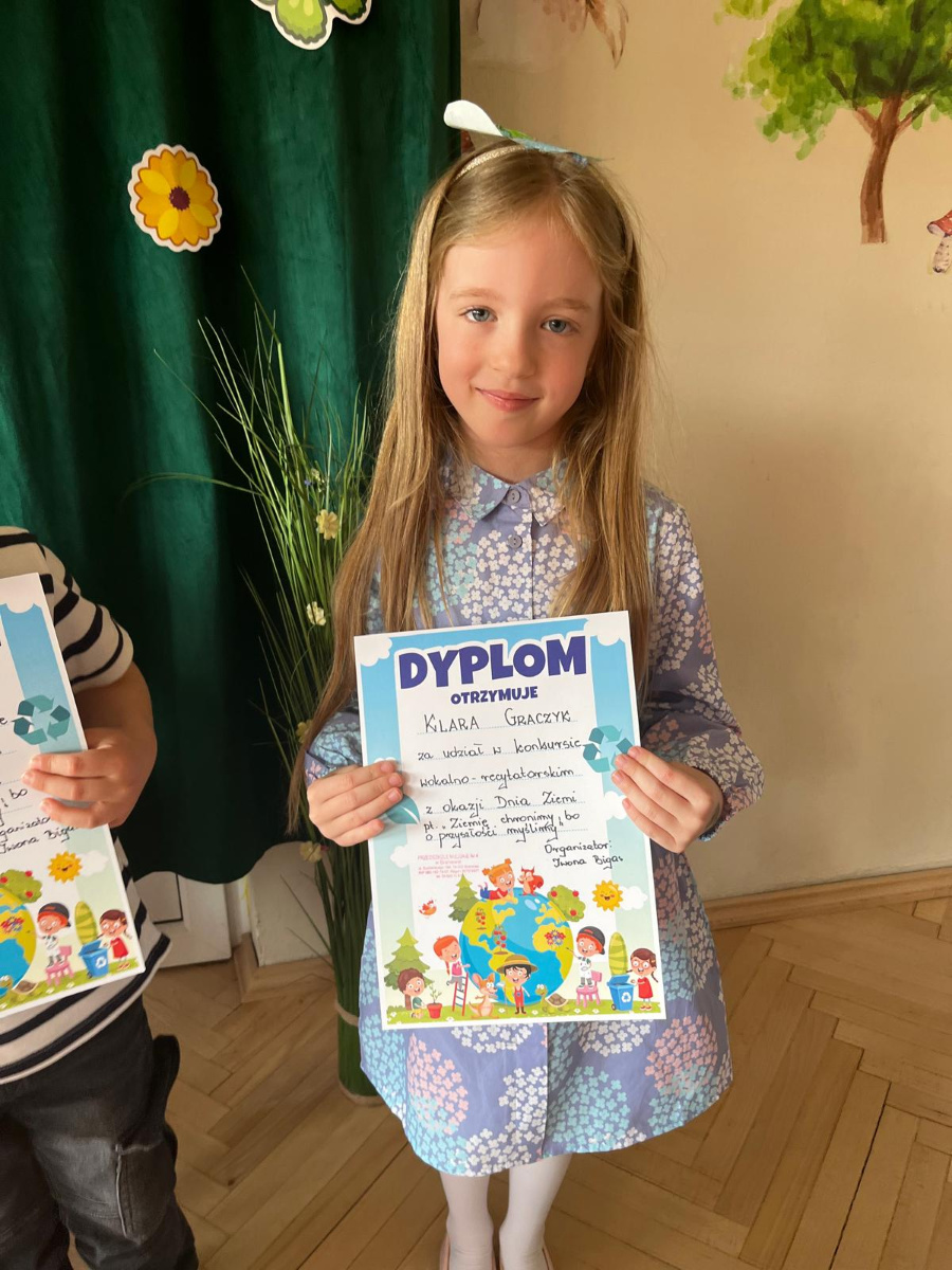 Dziewczynka w jasnoniebieskiej sukience w kwiaty trzyma w rękach dyplom. Dokument przyznany jest za udział w konkursie recytatorskim. Na dyplomie znajdują się kolorowe ilustracje dzieci 
