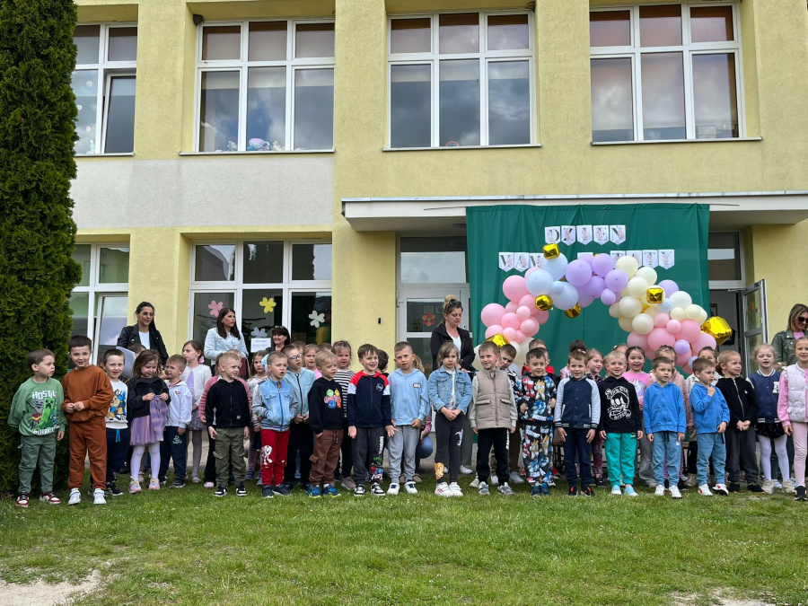 Grupa dzieci stoi przed żółtym budynkiem, tworząc szereg. Za nimi widać zieloną dekorację z napisem „Dzień Mamy i Taty” oraz kolorowy łuk z balonów w pastelowych barwach