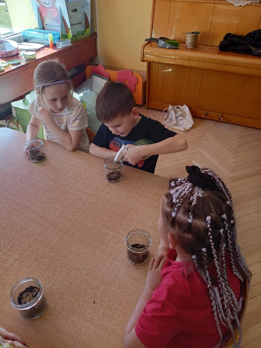 Troje dzieci siedzi przy stoliku i pracuje z małymi słoikami wypełnionymi ziemią. Chłopiec wsypuje do swojego słoika kolejną warstwę przy pomocy łyżeczki