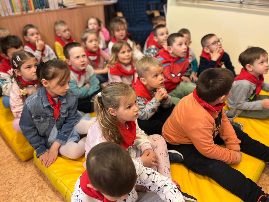 Grupa dzieci siedzi blisko siebie na żółtych matach patrząc w jednym kierunku. Większość przedszkolaków ma na sobie czerwone chusty na szyjach. W tle widać półki z książkami