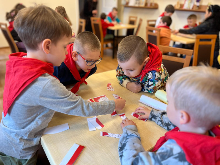 Grupa dzieci siedzią przy stoliku i układa puzzle z elementów w barwach biało czerwonych. W tle widać inne stoliki przy których również pracują dzieci