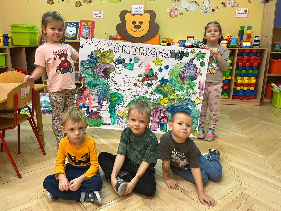 Grupa dzieci pozuje do zdjęcia trzymając dużą pokolorowaną planszę z napisem ANDRZEJKI. Na rysunku widoczna jest kolorowa ilustracja z gwiazdkami i magicznymi przedmiotami