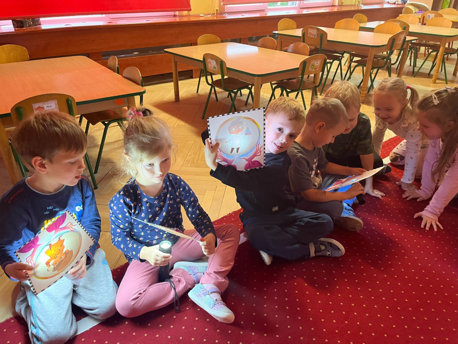 Grupa dzieci siedzi na czerwonym dywanie trzymając kolorowe kartki z rysunkami. Jedno z dzieci używa latarki oświetlając obrazek. W tle widoczna jest część sali z drewnianymi stołami i krzesłami