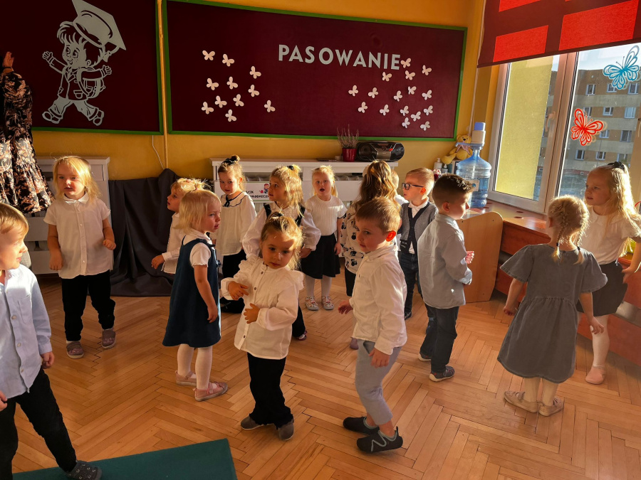 Dzieci ubrane w białe koszule i ciemne spodnie tańczą ze sobą. Na ścianie widoczny jest napis Pasowanie oraz dekoracja w postaci białych motylków