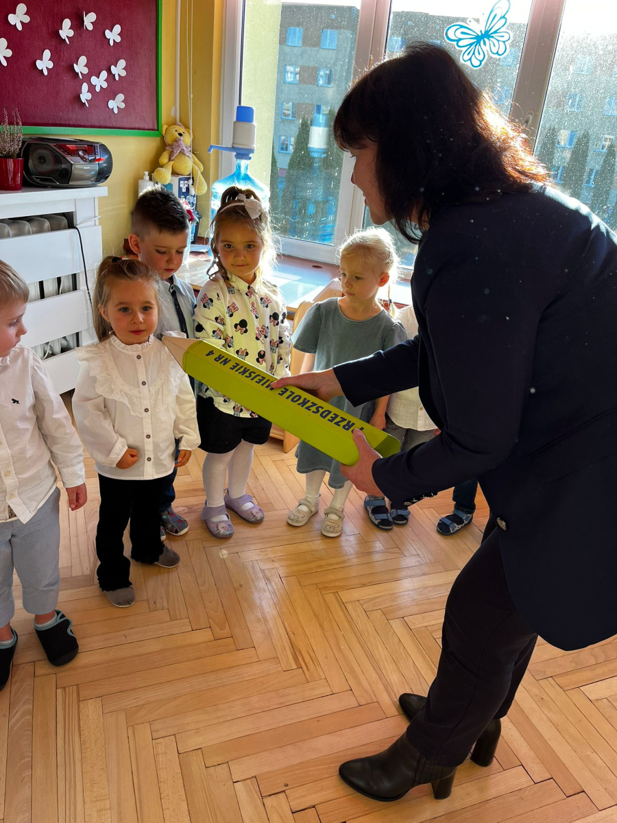 Grupa dzieci stoi w kolejce w sali przedszkolnej ubrane w białe koszule. Kobieta ubrana na czarno kładzie dziewczynce dużą kredkę na ramieniu