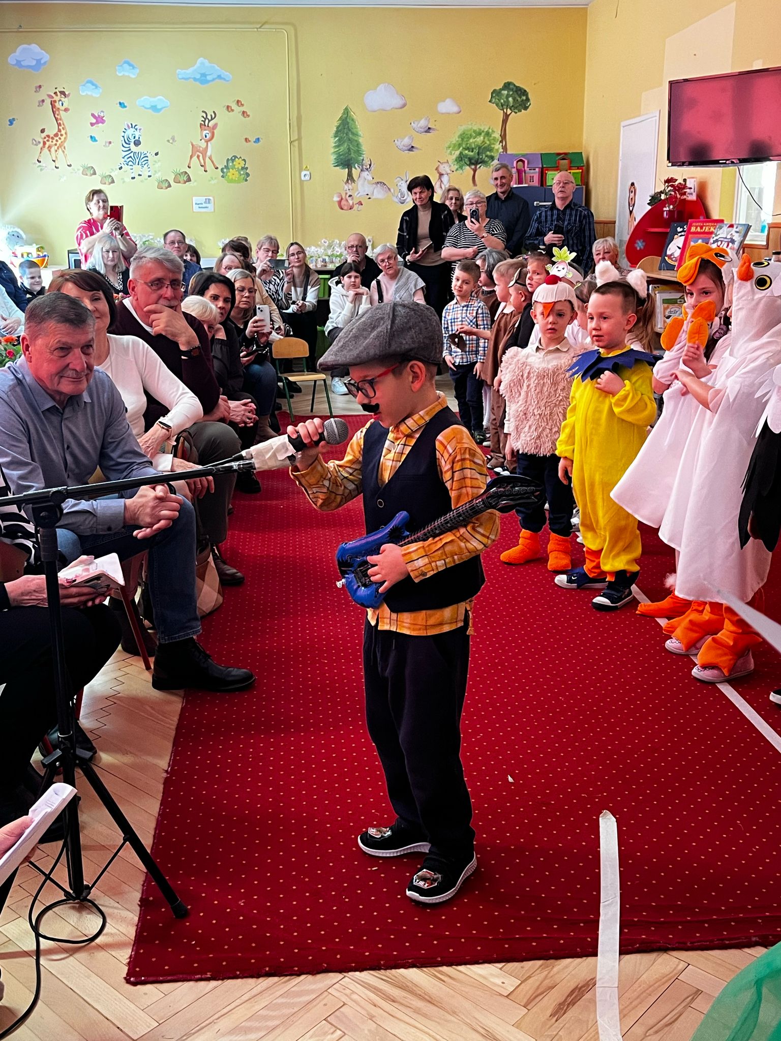 Chłopiec w przebraniu dziadka z czapką okularami i sztucznymi wąsami stoi na czerwonym dywanie trzymając w ręku mały instrument. Za nim ustawione są dzieci w kolorowych kostiumach