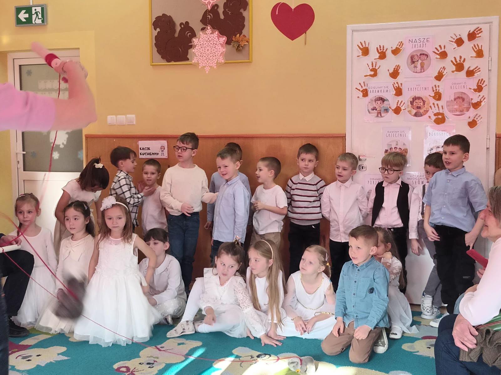 Grupa dzieci stoi i siedzi na dywanie. Dziewczynki ubrane są w białe sukienki a chłopcy w koszule i eleganckie spodnie. W tle na drzwiach widnieją kolorowe plakaty