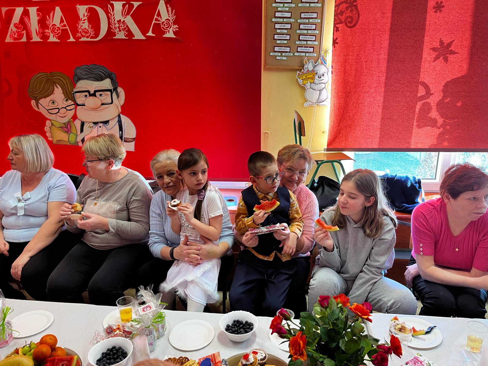 Grupa osób siedzi obok siebie na ławce. Dziewczynka w białej sukience siedzi na kolanach starszej kobiety, obok chłopiec w okularach trzymający kawałek arbuza