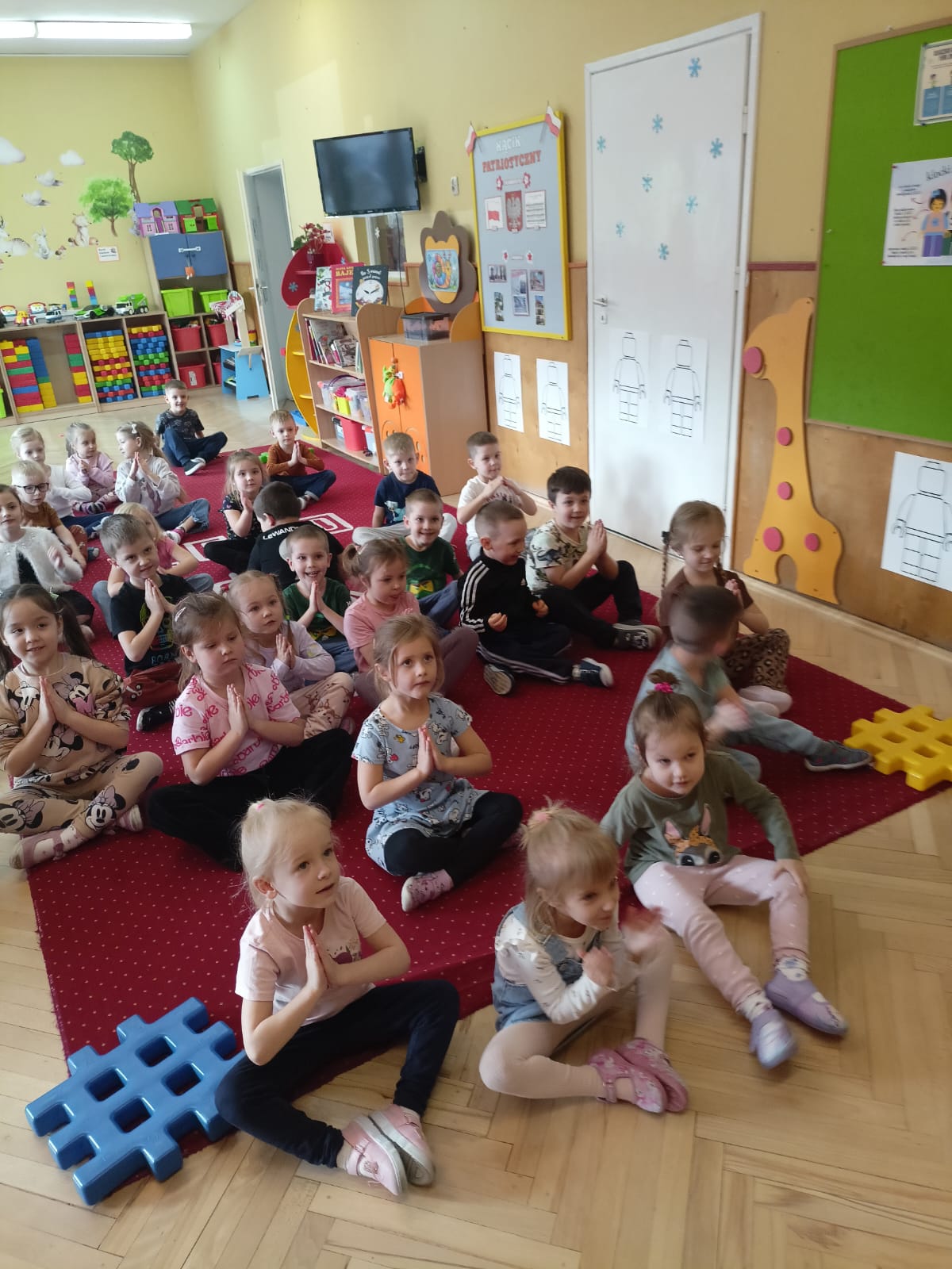 Grupa dzieci siedzi na czerwonym dywanie w sali pełnej kolorowych zabawek i mebli. Dzieci siedzą w rzędach część z nich trzyma złożone dłonie a inne patrzą przed siebie z uśmiechem