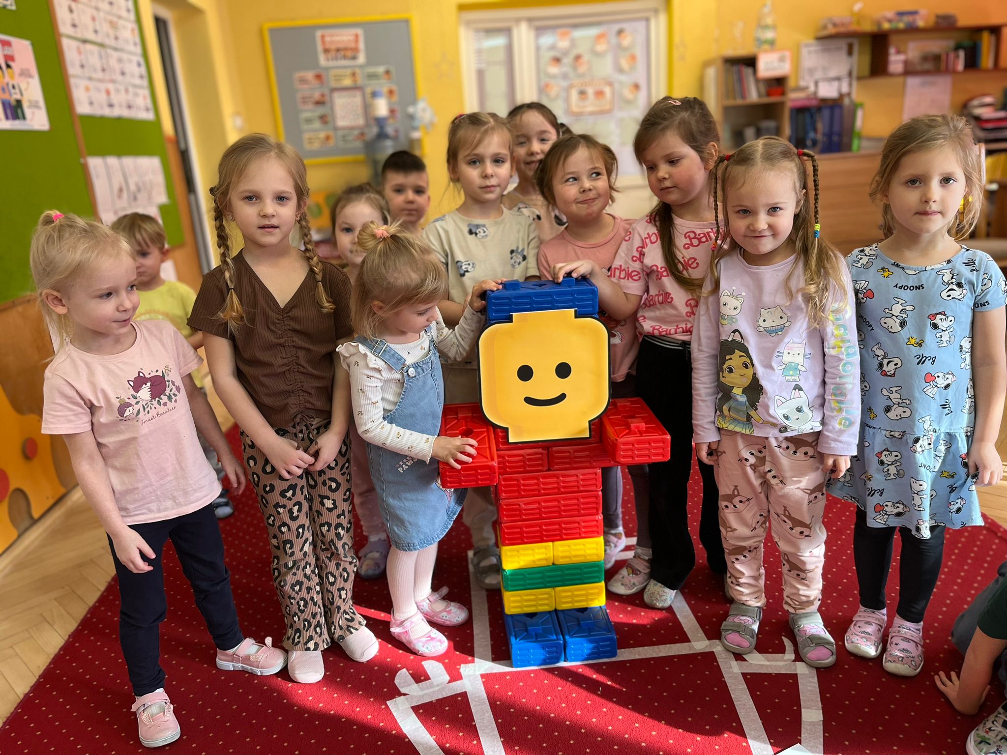 Grupa dzieci stoi wokół dużej kolorowej postaci zbudowanej z klocków. Postać ma żółtą głowę z uśmiechniętą buzią, czerwony tułów, niebieskie nogi oraz pas w kolorze zielonym i żółtym