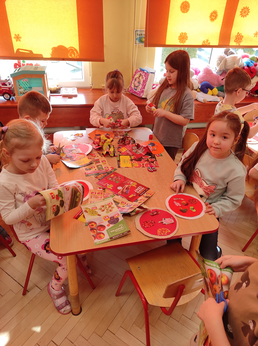 Dzieci siedzą przy stole i wykonują prace plastyczne z kolorowych wycinków gazet. Na stole leżą nożyczki kleje oraz wiele wyciętych obrazków przedstawiających jedzenie
