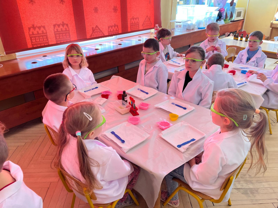 Dzieci ubrane w białe fartuchy i ochronne okulary siedzą przy stolikach przygotowanych do zajęć laboratoryjnych. Na stołach znajdują się tace, kolorowe naczynia, plastikowe łyżeczki oraz buteleczki