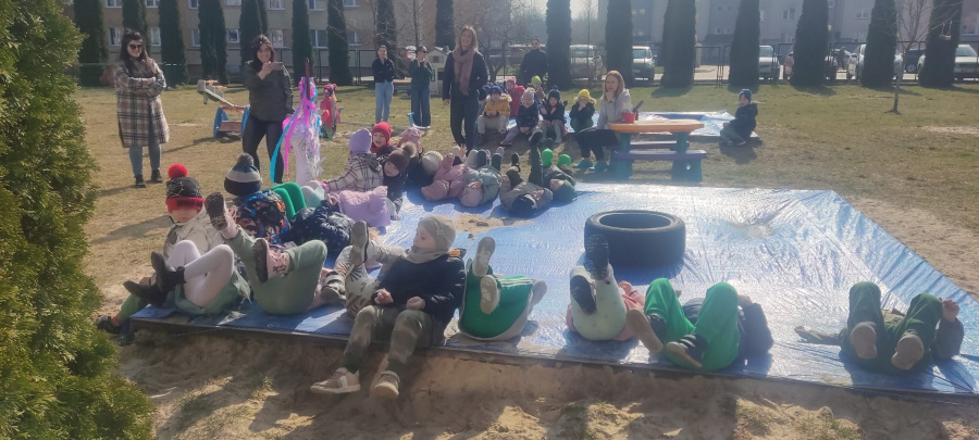 Dzieci leżą na plecach na dużej niebieskiej plandece rozłożonej na piaskownicy i unoszą nogi do góry. W tle stoją nauczycielki oraz inne dzieci obok widoczna jest kolorowa kukła ze słomy