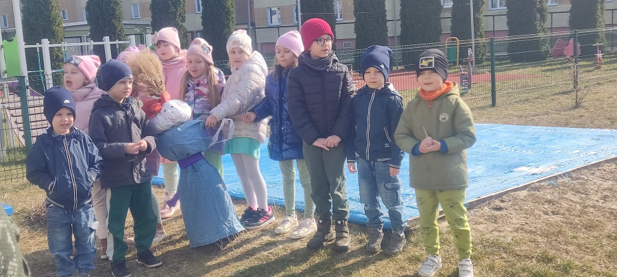 Dzieci stoją na świeżym powietrzu obok dużej kukły wykonanej ze słomy i ubranek. Dzieci mają na sobie ciepłe kurtki i czapki a w tle widać ogrodzenie placu zabaw oraz budynki