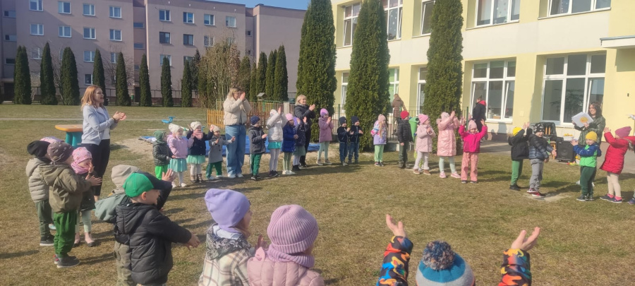 Dzieci stoją w dużym kole na trawniku przed budynkiem. Część z nich unosi ręce do góry inne klaszczą. Obok dzieci stoją nauczycielki. W tle widać wysokie iglaki oraz budynki mieszkalne