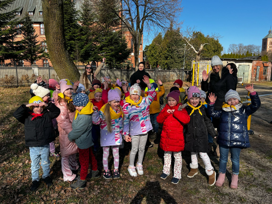 Grupa dzieci stoi razem w parku. Ubrane w kolorowe kurtki, czapki i szaliki, machają rękami do góry. Obok nich stoją nauczycielki. W tle widoczne są drzewa, ogrodzenie i ceglane budynki