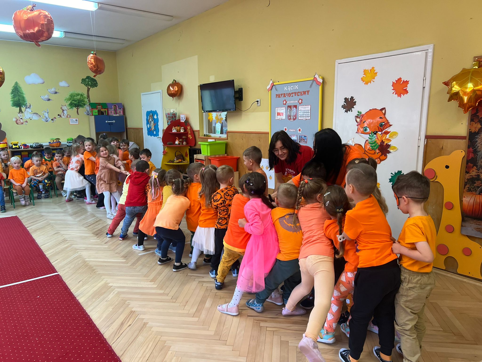 Dzieci przeciągają linę. Dwie grupy ubrane w pomarańczowe ubrania próbują przeciągnąć linę na swoją stronę. W tle widoczna dekoracja z jesiennymi motywami kolorowymi liśćmi dyniami 