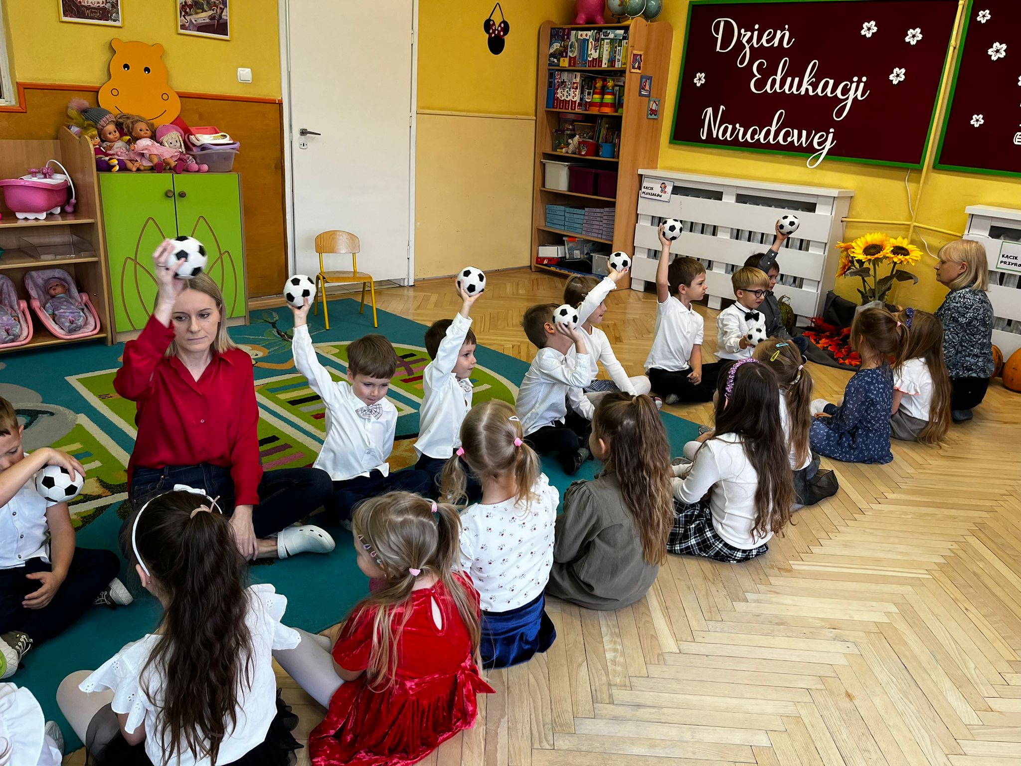 Dzieci siedzące naprzeciwko siebie na dywanie. Każde z dzieci trzyma w ręku piłkę w biało czarną kratę unosząc ją do góry. W tle widoczna dekoracja z napisem Dzień Edukacji Narodowej 