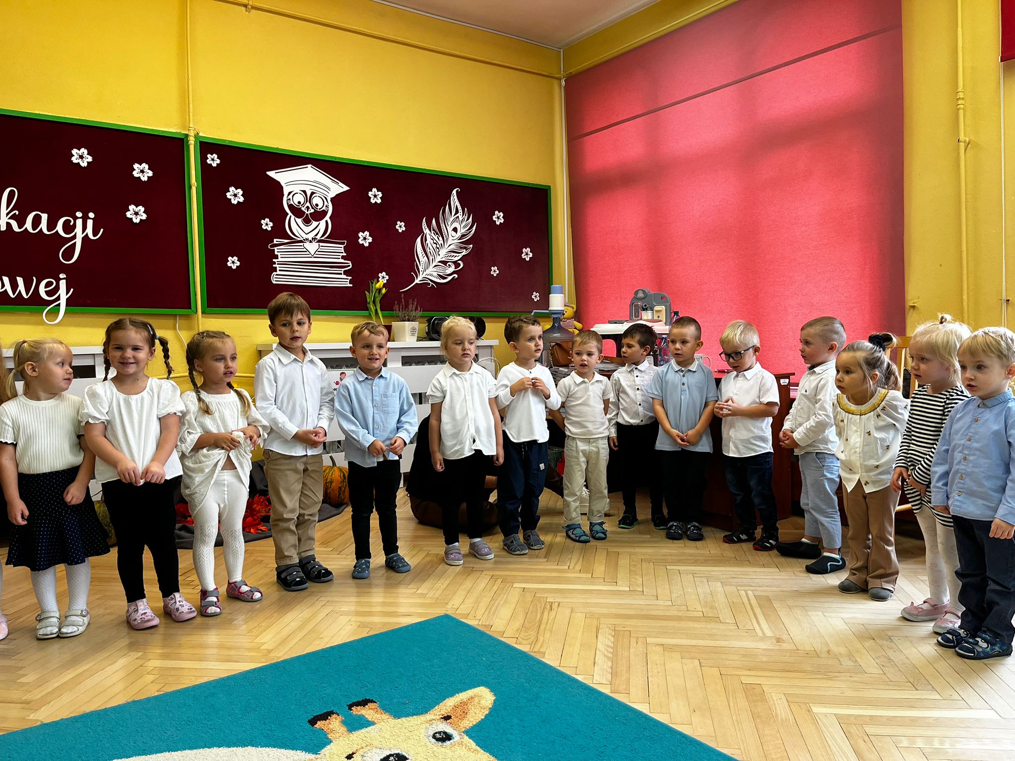 Grupa dzieci stoi w szeregu w sali większość dzieci ma białe bluzki i ciemne spodnie lub spódniczki. W tle dekoracja sowa w birecie książki oraz pióro otoczone kwiatkami. 