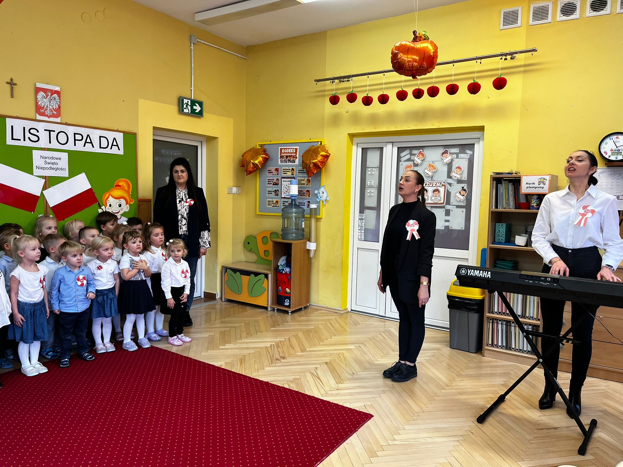 Grupa dzieci ubranych w białe koszule stoi na czerwonym dywanie. Z prawej strony dwie kobiety jedna śpiewa druga gra na klawiszach Yamaha
