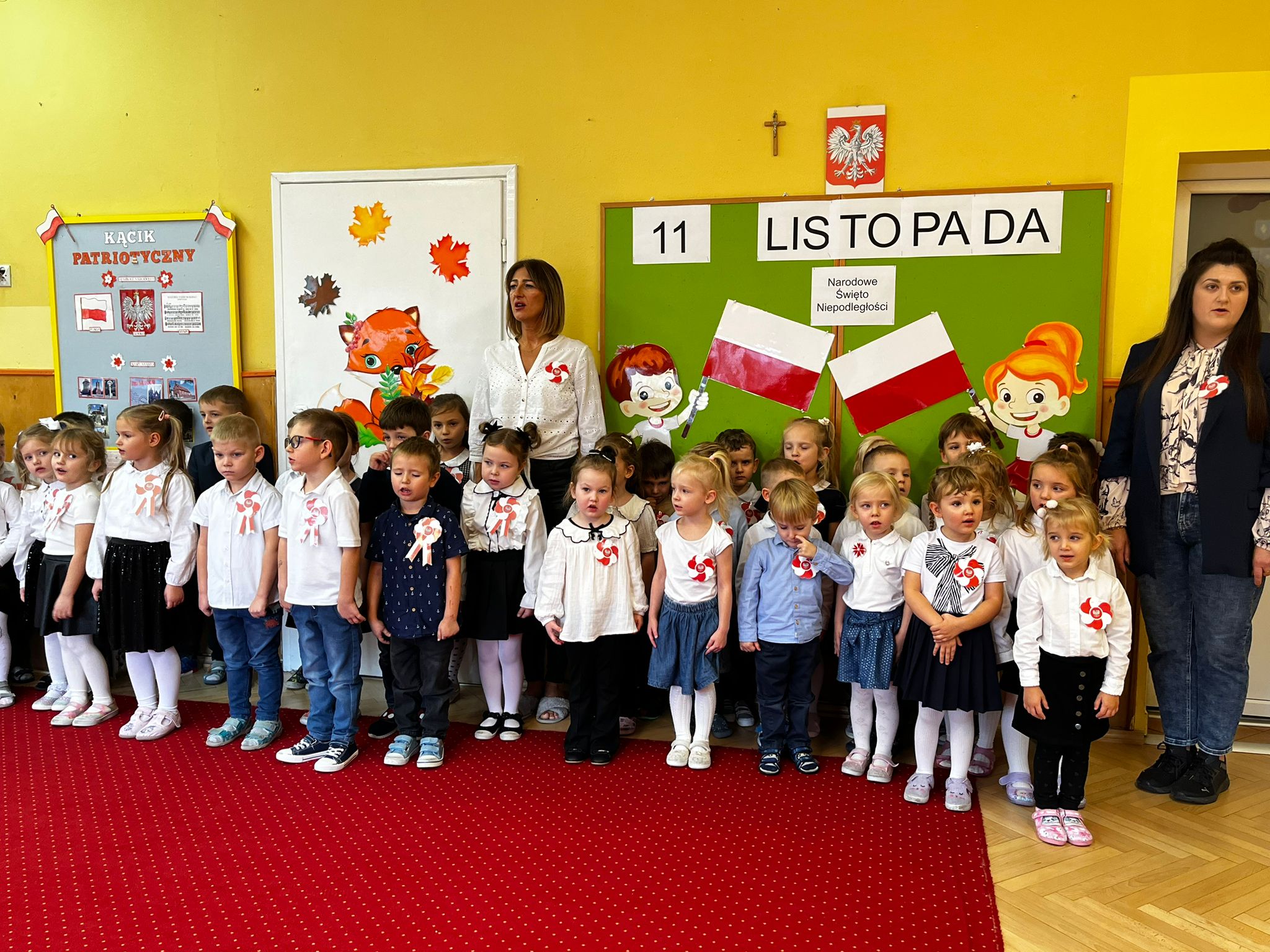 Grupa dzieci ubranych w biało czarne stroje stoi na czerwonym dywanie. W tle widoczna jest ilustracja dzieci trzymających polskie flagi