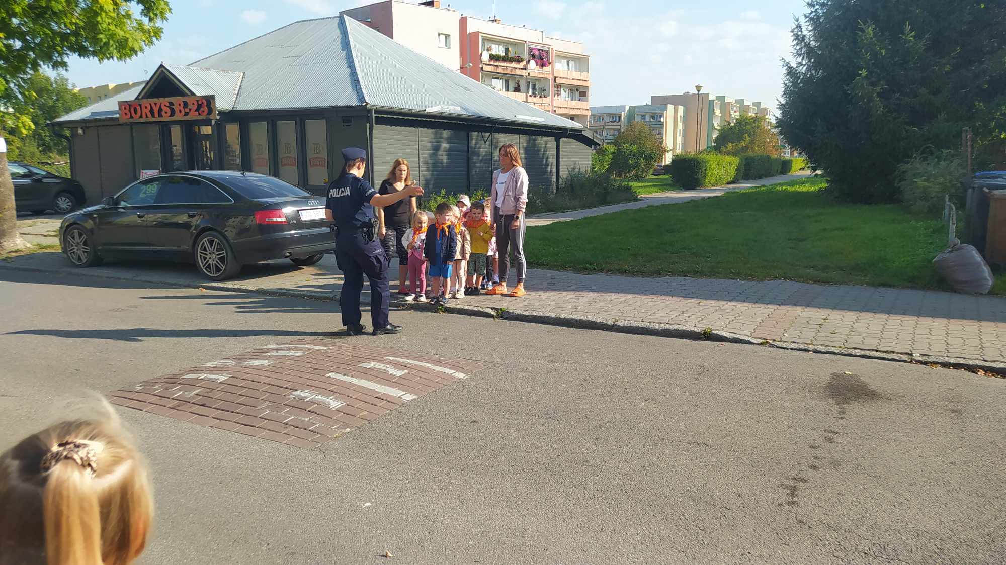 grupa dzieci stojących na chodniku przed nimi na trawie stoji policjantka