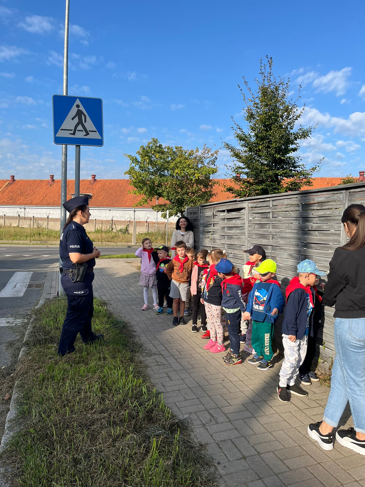 grupa dzieci stojąca na chodniku przed nimi stoji policjantka po lewej stornie przejście dla pieszych
