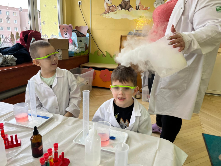 Dwóch chłopców w białych fartuchach i ochronnych okularach siedzi przy stole. Nauczycielka w fartuchu laboratoryjnym trzyma kolbę, z której wydobywa się gęsta para przypominająca dym