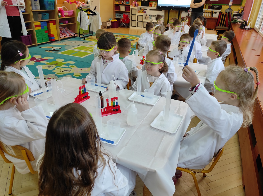 Dzieci ubrane w białe fartuchy i ochronne okulary siedzą przy stołach. Przed nimi znajdują się tacki z naczyniami laboratoryjnymi pipetami zlewkami probówkami i stojakami