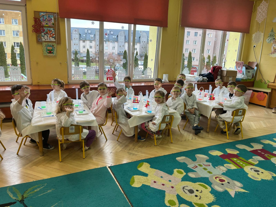 Grupa przedszkolaków w białych fartuchach i okularach ochronnych siedzi przy trzech stołach ustawionych w sali z dużymi oknami