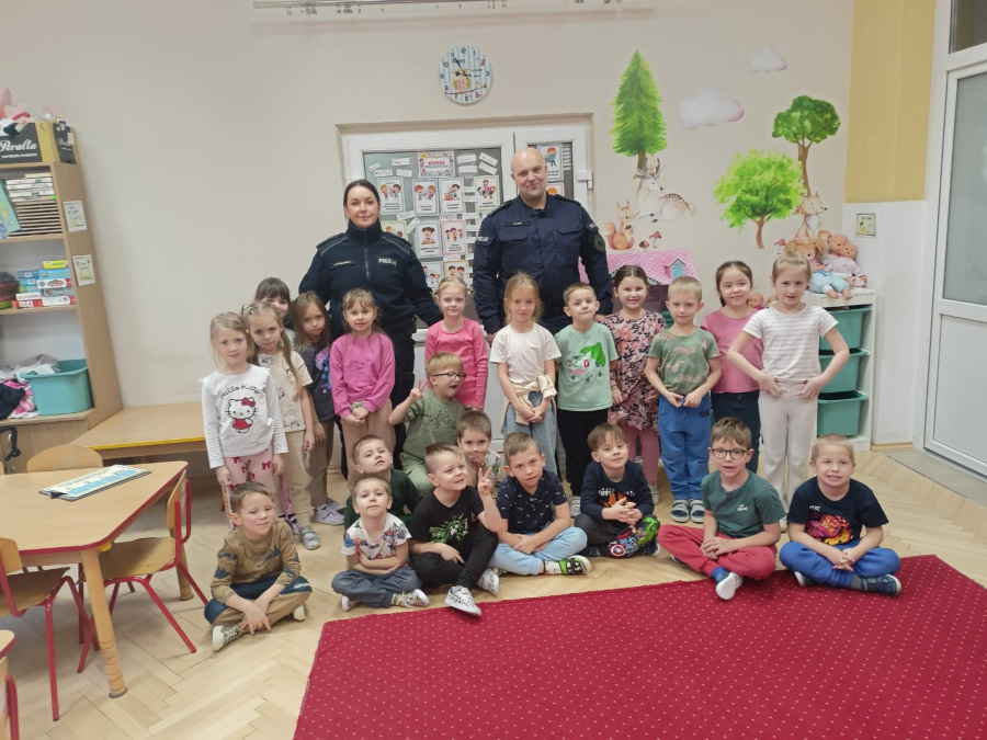 Dzieci pozują do zdjęcia grupowego z dwójką policjantów w sali przedszkolnej. Funkcjonariusze stoją z tyłu