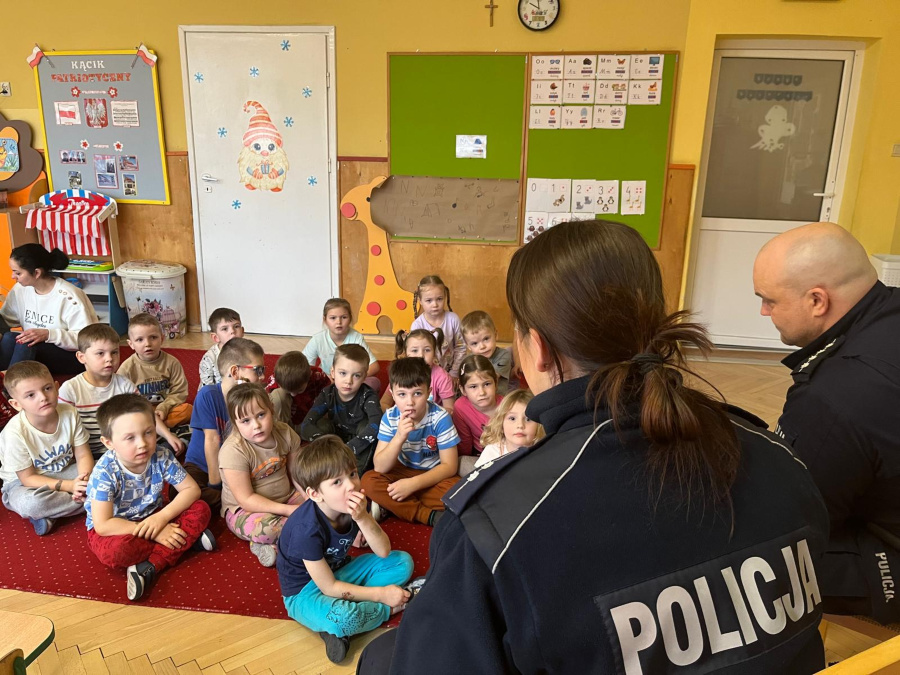 Dzieci siedzą na czerwonym dywanie i słuchają dwójki policjantów, którzy siedzą tyłem do obiektywu. Na plecach mundurów widnieje napis Policja