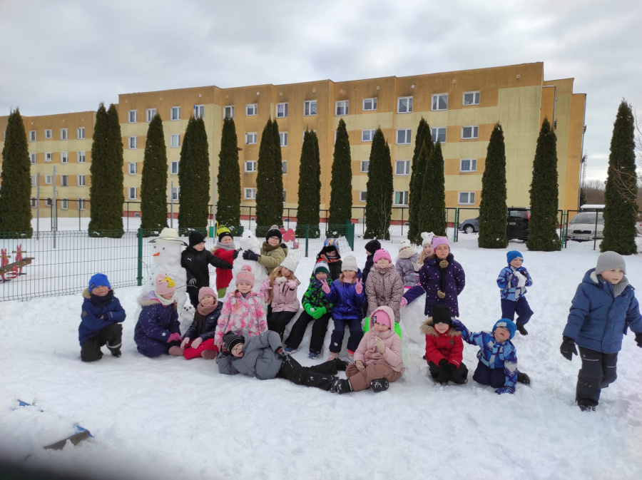 Grupa dzieci pozuje do wspólnego zdjęcia na śnieżnym placu zabaw. W tle stoją dwa ulepione bałwanki jeden z nich ma kapelusz. Dzieci są ubrane w zimowe kombinezony