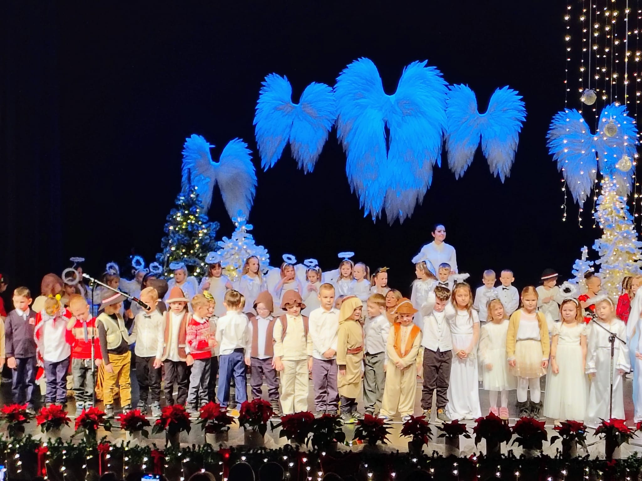 Grupa dzieci stoi na scenie podczas występu świątecznego. Dzieci ubrane w stroje aniołów, pastuszków i odświętne ubrania. Nad nimi dekoracje skrzydeł, oświetlone choinki i świąteczne lampki