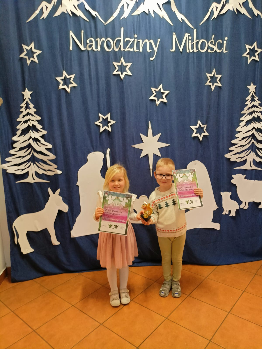 Dwoje dzieci stoi przed granatową dekoracją świąteczną z napisem Narodziny Miłości. Trzymają dyplomy jedno z nich dodatkowo figurkę. W tle gwiazdy choinki i sylwetki postaci bożonarodzeniowych